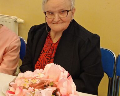 Zbliżenie. Seniorka w ciemnej marynarce i i czerwonej bluzce siedzi przy stole, kobieta uśmiecha się i pozuje do zdjęcia.