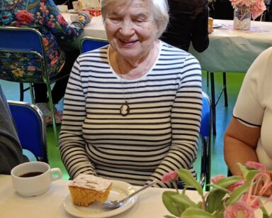 Zbliżenie. Uśmiechnięta seniorka w bluzce w paski siedzi przy stole i patrzy na ciasto. W tle grupa ludzi przy stole.