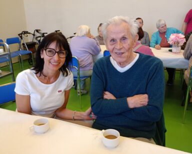 Zbliżenie. Przy stole młoda kobieta w jasnej koszulce i senior ze złożonymi na piersi rękoma. Para pozuje do zdjęcia.