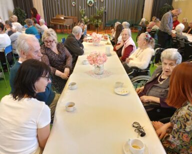 Sala. Przy stołach siedzi grupa osób, seniorzy, opiekunowie i młodzi ludzie. Na stolach talerzyki i filiżanki oraz kwiaty.