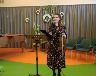 Zbliżenie. Na scenie młoda kobieta w sukience w kwiaty, w ręku trzyma skrzypce. Przed nią mikrofon i stojak na nuty.