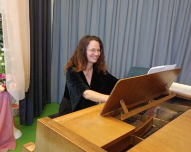 Zbliżenie. Kobieta siedzi przy fortepianie, uśmiecha się. Jest w okularach, ma długie kręcone włosy i czarną bluzkę.