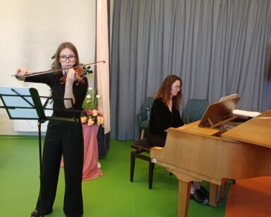 Scena. Przy stojaku z nutami stoi młoda kobieta i gra na skrzypcach. W tle kobieta gra na fortepianie.