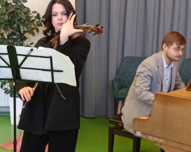 Scena. Przy stojaku z nutami stoi młoda kobieta i gra na skrzypcach. W tle mężczyzna w jasnym garniturze gra na fortepianie.