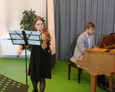 Scena. Przy stojaku z nutami stoi młoda kobieta i gra na skrzypcach. W tle mężczyzna w jasnym garniturze gra na fortepianie.