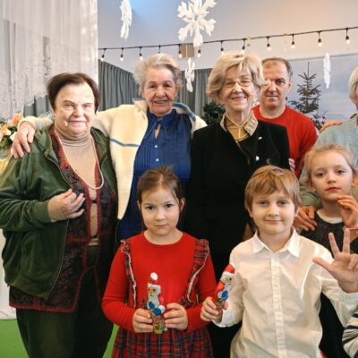 Zbliżenie. Czworo seniorów i troje maluchów pozują do wspólnego zdjęcia. W tle z sufitu zwisają śnieżynki.