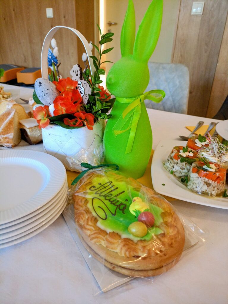 Zbliżenie. Stół. Na stole zielony zajączek, koszyczek z ozdobą wielkanocną, ciasta, galaretki i talerze.