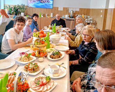 Sala. Przy stole zastawionym wielkanocnym jedzeniem i ozdobami siedzi grupa osób. Wolontariusze pozują do zdjęcia.
