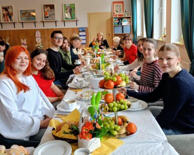 Sala. Przy stole zastawionym wielkanocnym jedzeniem i ozdobami siedzi grupa osób. Wolontariusze pozują do zdjęcia.