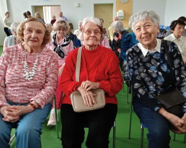 Zbliżenie. Trzy uśmiechnięte seniorki pozują do zdjęcia. W tle grupa ludzi.