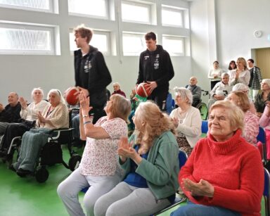 Sala. Grupa seniorów siedzących na krzesłach, oklaskami wita dwóch wysokich koszykarzy. Mężczyźni trzymają w rękach piłki.