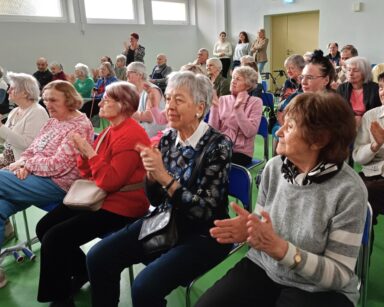 Sala. Grupa seniorów siedzących na krzesłach, oklaskami wita gości. W tle trzy kobiety stoją przy ścianie.
