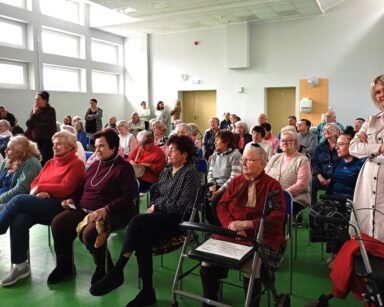 Sala. Grupa seniorów słucha wywiadu młodych koszykarzy. W tle kilka osób stoi i ogląda wywiad z zaciekawieniem.
