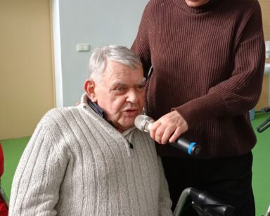 Zbliżenie. Senior w szarym swetrze mówi do mikrofonu , który trzyma kobieta w ciemnych włosach i brązowym swetrze.