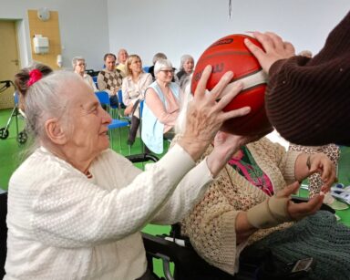 Zbliżenie. Seniorka w jasnym swetrze trzyma piłkę do koszykówki. Kobita uśmiecha się.