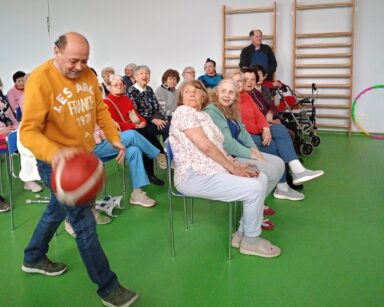 Sala. Senior w żółtej bluzie kozłuje piłkę do koszykówki. W tle grupa seniorów ogląda jego występ.