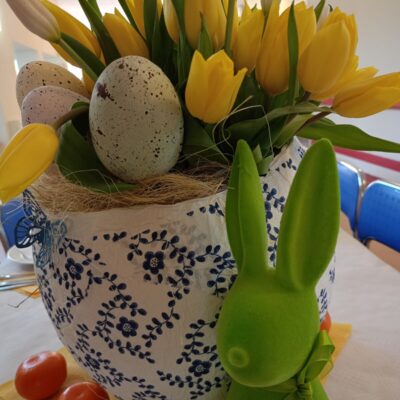 Zbliżenie. Jajko wyklejanka jako wazon. w wazonie żółte tulipany. Obok figurka zielonego zająca i rozłożone mandarynki.