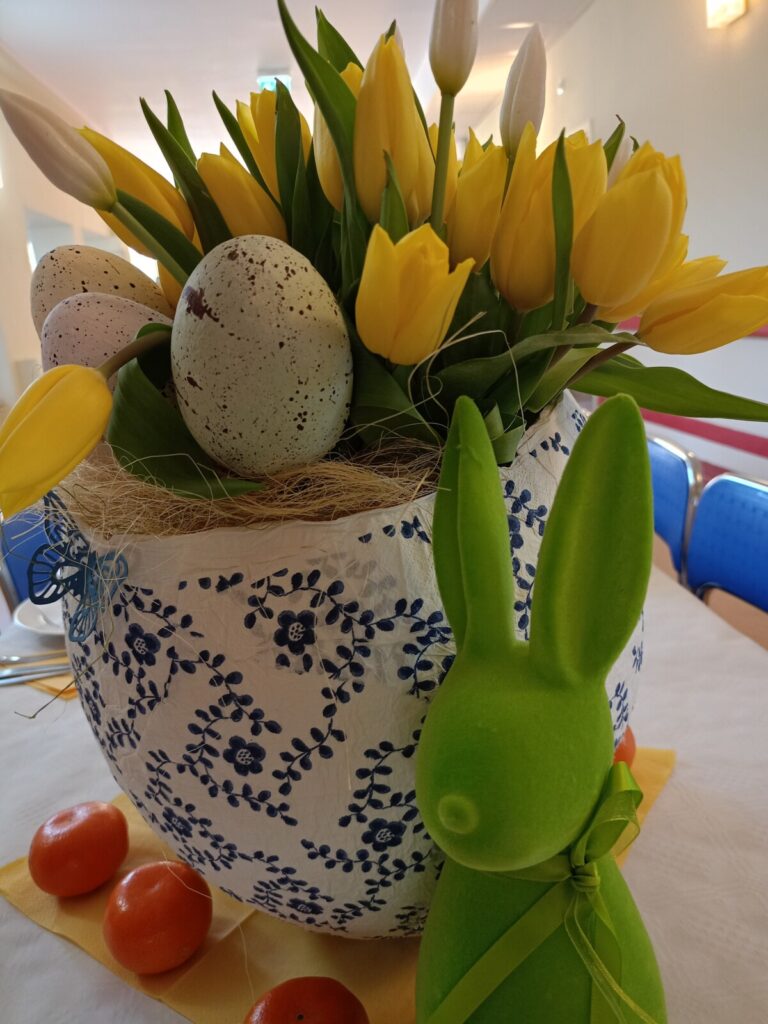 Zbliżenie. Jajko wyklejanka jako wazon. w wazonie żółte tulipany. Obok figurka zielonego zająca i rozłożone mandarynki.