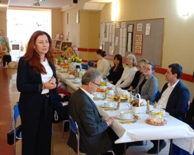 Sala. Przy stołach siedzi grupa osób. Kobieta w rudych włosach, w ciemnym garniturze przemawia do grona osób.