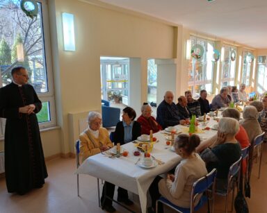 Sala. Przy stołach siedzi grupa osób. Ksiądz przemawia do grona osób. Na stołach zastawa, chleb, owoce.