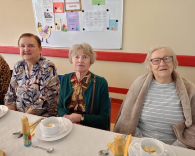 Zbliżenie. Trzy uśmiechnięte seniorki pozują do zdjęcia. Kobiety siedzą przy zastawionym stole.