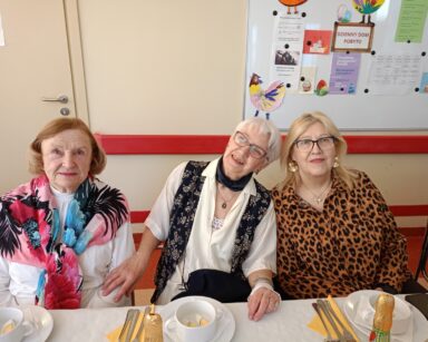 Zbliżenie. Trzy uśmiechnięte seniorki pozują do zdjęcia. Kobiety siedzą przy zastawionym stole.