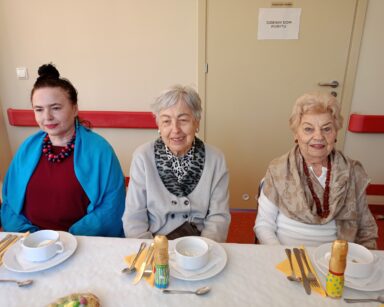 Zbliżenie. Trzy uśmiechnięte seniorki pozują do zdjęcia. Kobiety siedzą przy zastawionym stole.