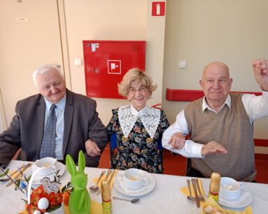Zbliżenie. Troje uśmiechniętych seniorów pozuje do zdjęcia. Seniorzy siedzą przy zastawionym stole.