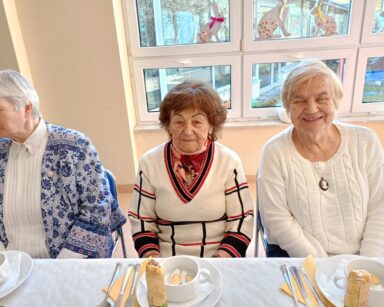 Zbliżenie. Trzy uśmiechnięte seniorki pozują do zdjęcia. Kobiety siedzą przy zastawionym stole.