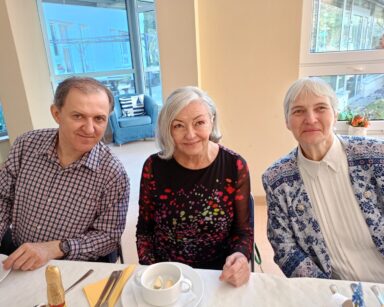 Zbliżenie. Troje uśmiechniętych seniorów pozuje do zdjęcia. Seniorzy siedzą przy zastawionym stole.