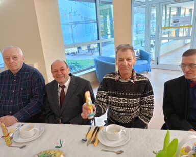 Zbliżenie. Trzech uśmiechniętych seniorów pozuje do zdjęcia. Seniorzy siedzą przy zastawionym stole.