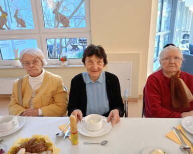 Zbliżenie. Trzy uśmiechnięte seniorki pozują do zdjęcia. Kobiety siedzą przy zastawionym stole.