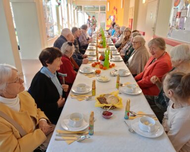 Sala. Długi stół, zastawiony wielkanocnym jedzeniem i dekoracjami. Przy stole siedzą seniorzy.