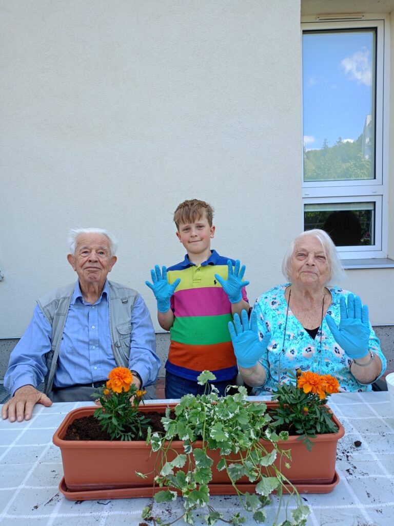 Zbliżenie. Para seniorów i chłopiec siedzą przy stole. Na stole stoi donica z kwiatami. Seniorka i chłopiec mają rękawice.