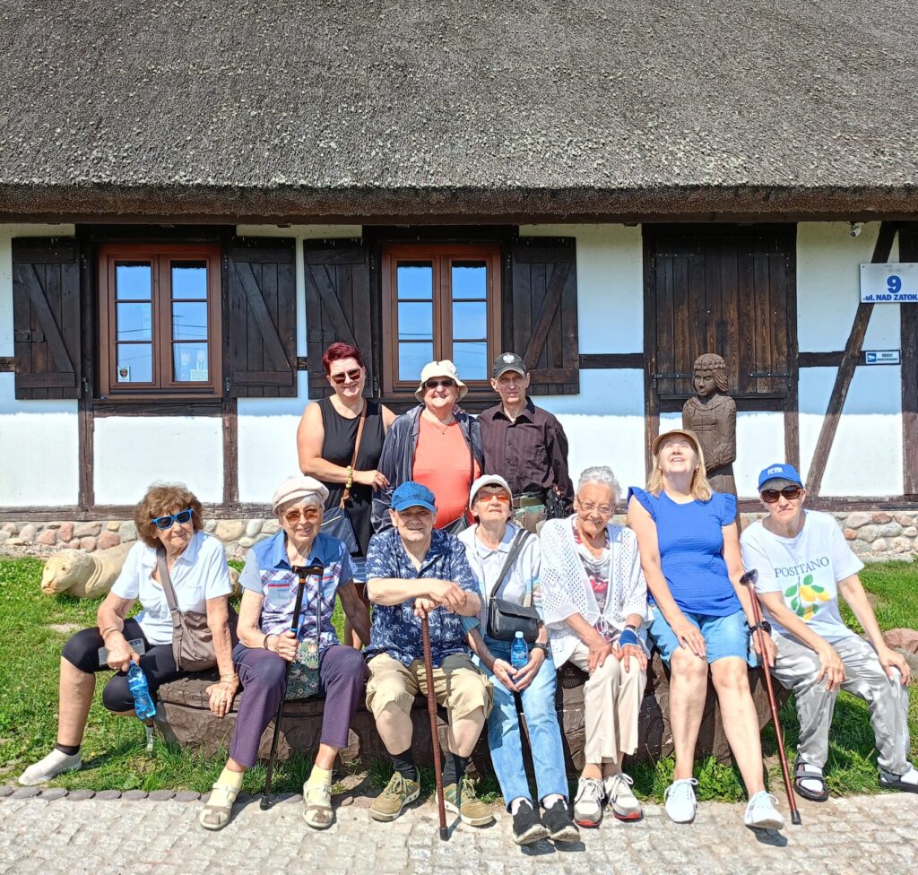 Zbliżenie. Grupa seniorów z DDP i wolontariuszka opiekunka pozują do zdjęcia na tle skansenu.